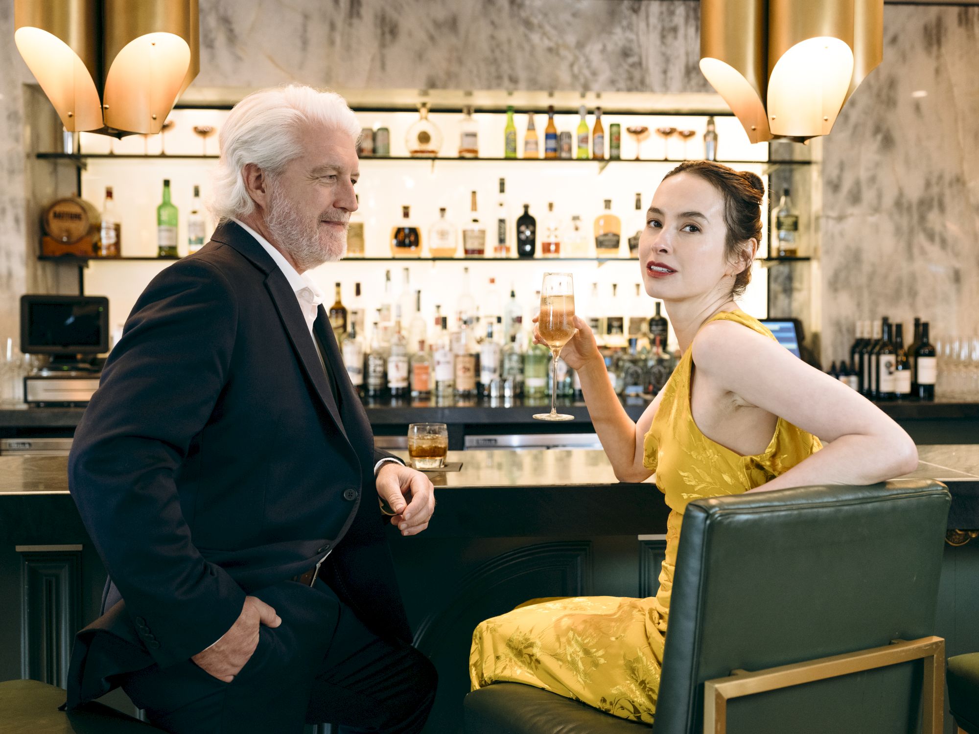 A man and a woman are sitting at a bar, engaged in conversation. The woman is holding a drink in her hand, while the man is smiling at her.