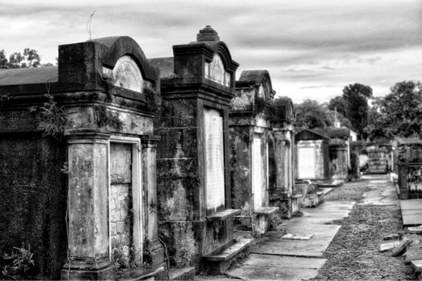 The image shows a black and white photo of an above-ground cemetery with weathered tombs and a cloudy sky.