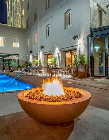 The image shows a luxurious outdoor area with a lit fire pit, a swimming pool, and seating, adjacent to a modern building at night.