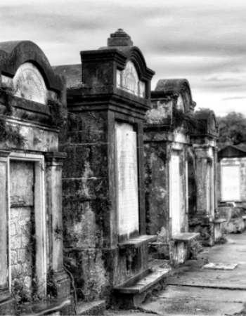 The image shows an old, atmospheric cemetery with stone tombs lined up along a path under a cloudy sky, captured in black and white.