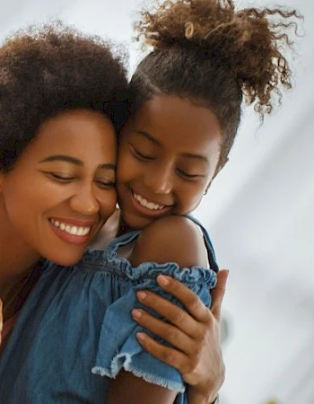 A woman and a girl are hugging and smiling on a couch, holding a bouquet of pink roses wrapped in brown paper.