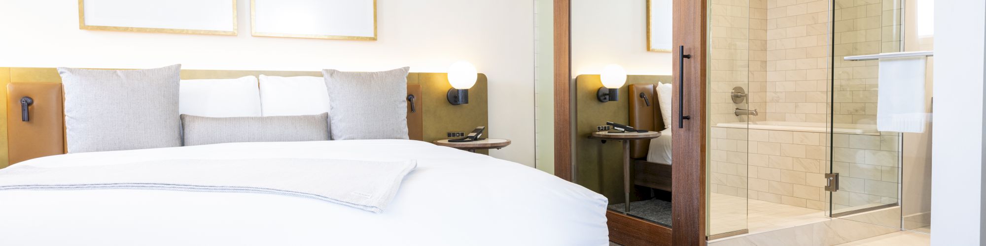 This image shows a modern hotel room with a large bed, two framed pictures on the wall, a bench at the foot of the bed, and an open glass shower area.