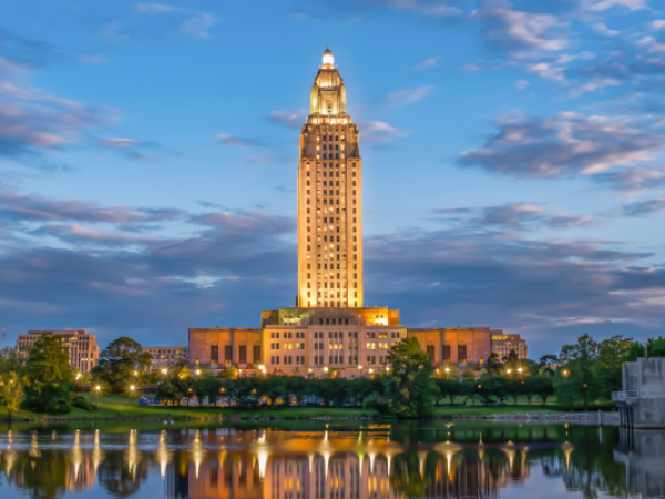 A tall, illuminated building with a reflective body of water in the foreground and a partly cloudy sky in the background.