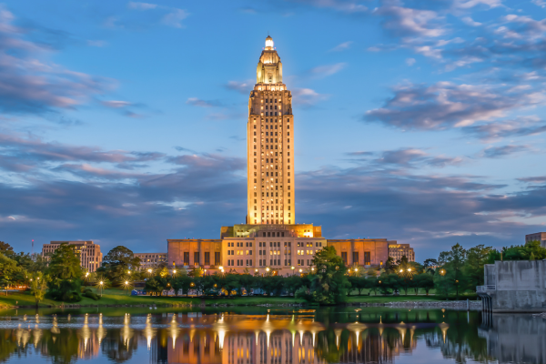 A tall, illuminated building with a reflective body of water in the foreground and a partly cloudy sky in the background.