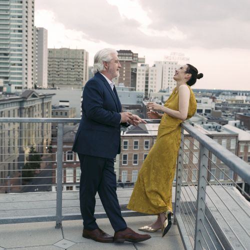 Two people are standing on a rooftop, engaged in conversation, with a cityscape view in the background.