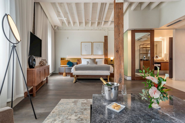 A modern bedroom features a bed, stylish furniture, a floor lamp, flowers, and a table with a champagne bucket in a spacious setting.