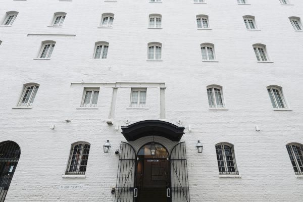 The image shows a white brick building with multiple windows and a central entrance with a black gate.