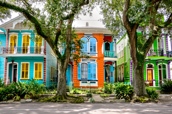 Colorful houses with vibrant facades stand behind large trees and greenery, creating a lively and picturesque street scene.