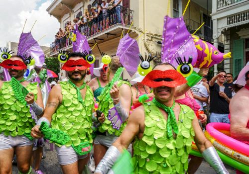 Costumed parade-goers in green leaf outfits and red mustache masks cheer on a street while colorful fish-shaped floats hang overhead.