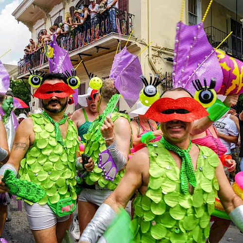 Costumed parade-goers in green leaf outfits and red mustache masks cheer on a street while colorful fish-shaped floats hang overhead.