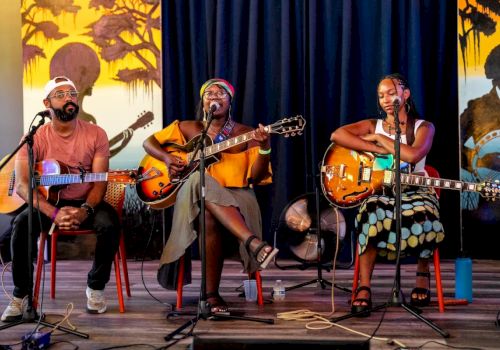 Three musicians perform on a stage with guitars and microphones, including a singer and two guitarists under spotlighted curtains.