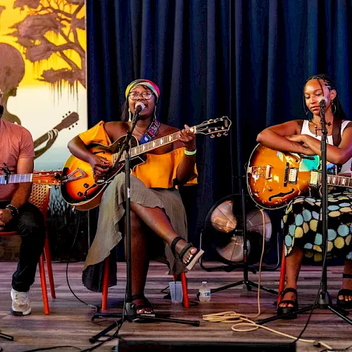 Three musicians perform on a stage with guitars and microphones, including a singer and two guitarists under spotlighted curtains.