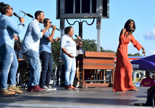 An outdoor stage where several people sing into microphones while a woman in a flowing orange dress performs.