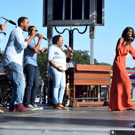 An outdoor stage where several people sing into microphones while a woman in a flowing orange dress performs.