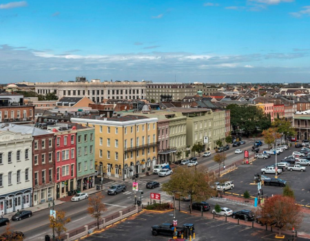 The image shows a cityscape with colorful buildings, a parking lot, cars, and a distant church steeple under a partly cloudy sky.