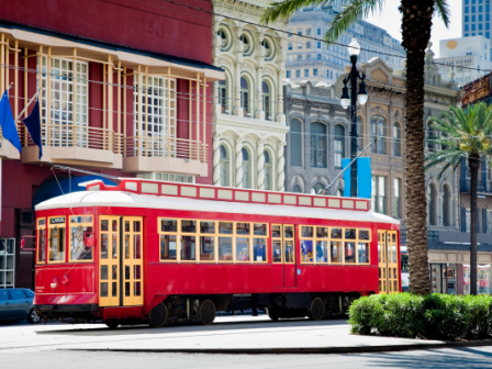 A vibrant red streetcar travels through an urban setting with palm trees, historic buildings, and clear skies.
