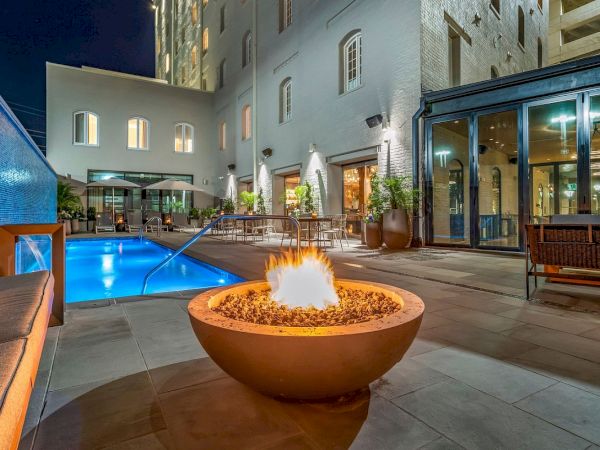 The image shows an outdoor patio with a fire pit and a swimming pool, surrounded by a modern building at night.