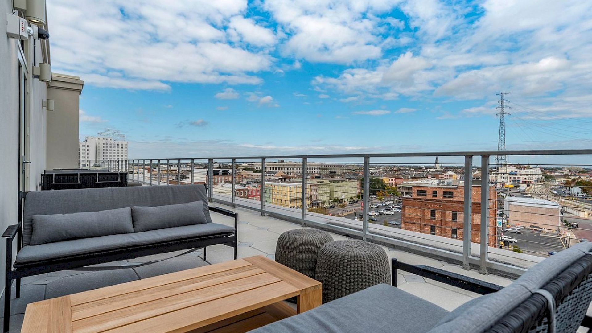 A rooftop patio with gray sectional seating, a wooden coffee table, and a panoramic city view under a blue sky with scattered clouds.