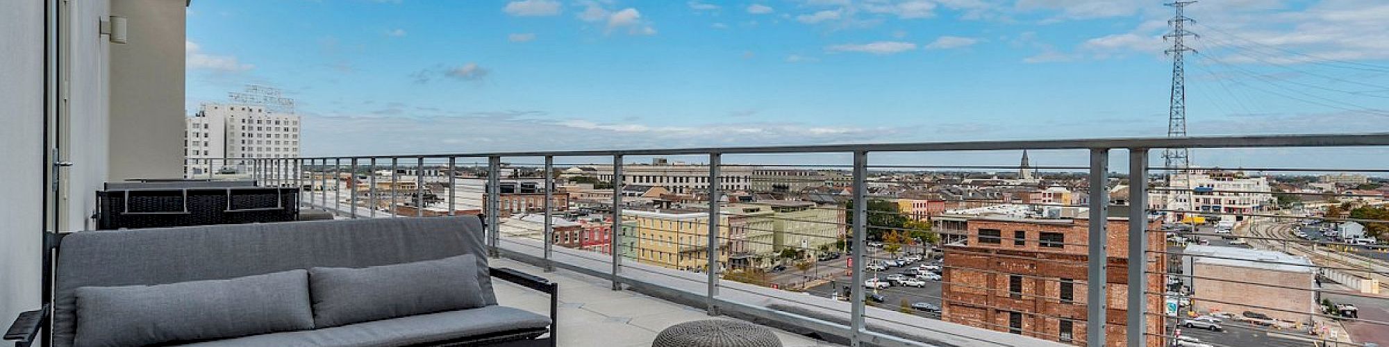 A rooftop patio with gray sectional seating, a wooden coffee table, and a panoramic city view under a blue sky with scattered clouds.