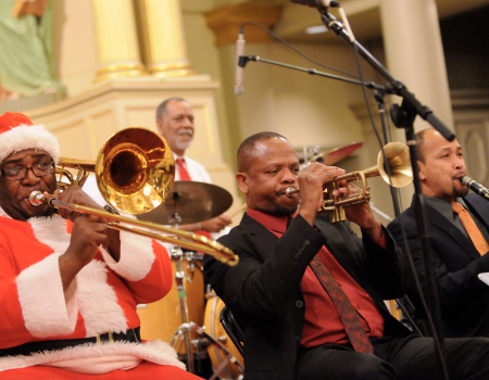 A festive band performs on stage, including a musician dressed as Santa Claus playing a trombone, with trumpets and drums in the background.