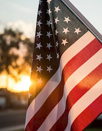 An American flag flutters in the sunset, rays glinting through the fabric as stars and stripes wave proudly in the breeze.