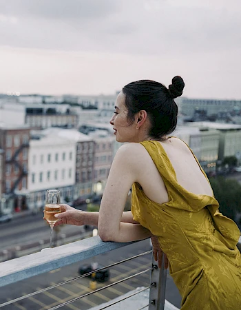 A woman in a yellow dress leans on a balcony railing, sipping wine and gazing over a city street and rooftops at dusk.