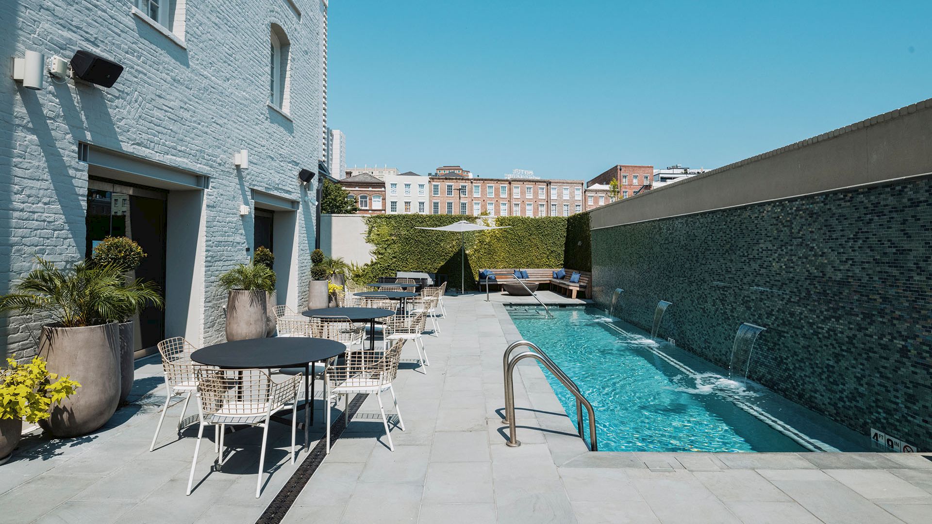 A modern pool area with loungers and round tables, bordered by white buildings on the left and a long blue-tiled pool on the right, sunny sky.