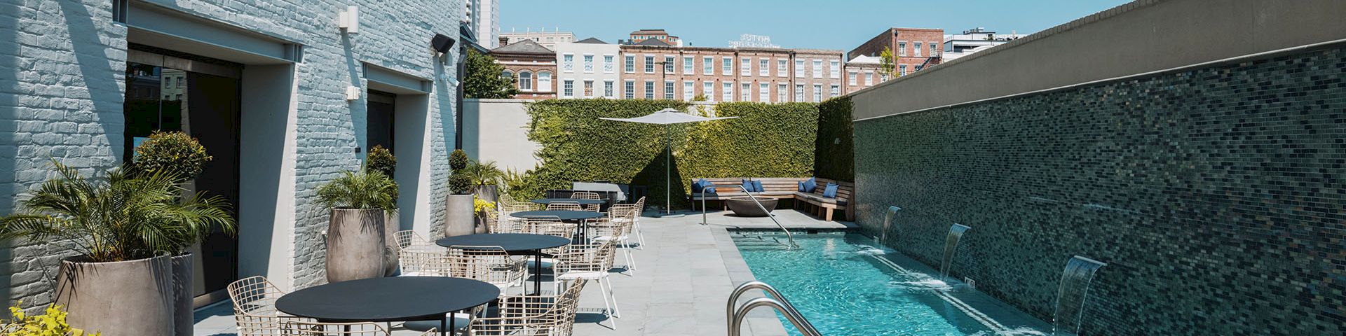 A modern pool area with loungers and round tables, bordered by white buildings on the left and a long blue-tiled pool on the right, sunny sky.