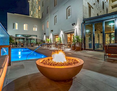 A night-time hotel courtyard with a glowing fire pit, blue pool, modern seating, and glass-walled lounge area. Ends with a period.
