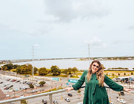 A woman in a green dress stands on a balcony overlooking a parking lot and water, smiling with arms outstretched at a sunny day.