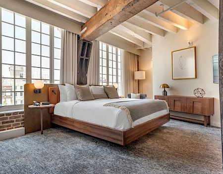 A stylish loft-style hotel room with a large bed, brick accent wall, wooden furniture, and big windows letting in natural light.