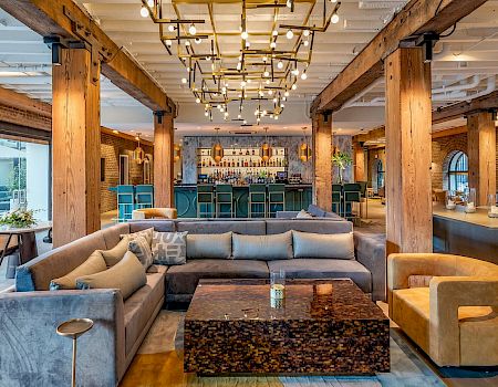 A modern, industrial-style lounge with plush gray and yellow seating, a dark marble coffee table, warm wooden beams, and a bar area in the background.