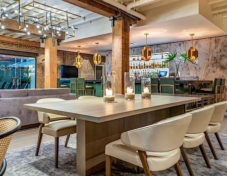 A modern, stylish restaurant with a large rectangular table in the foreground, white chairs, warm lighting, exposed brick, and a bar area in the background.