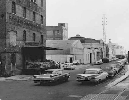 A black-and-white street scene with vintage cars parked along a curb, old brick buildings, warehouses, and a railroad track in the foreground.