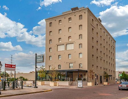 A tall beige brick building on a corner with shops at street level, under a blue sky with clouds.