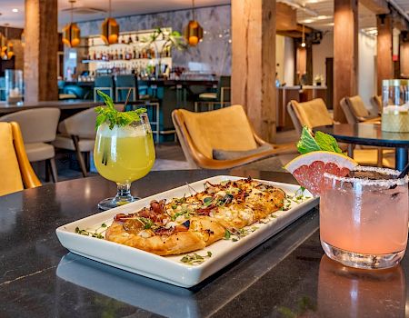 A colorful bar-restaurant scene with a flatbread pizza on a plate and two vibrant cocktails on a dark table, inviting and chic.