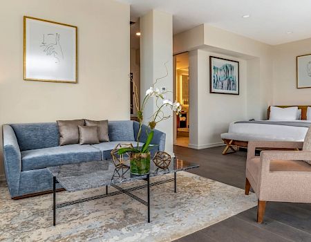 A stylish hotel suite with a living area and a bedroom, featuring a blue sofa, glass coffee table, and a cozy seating setup.
