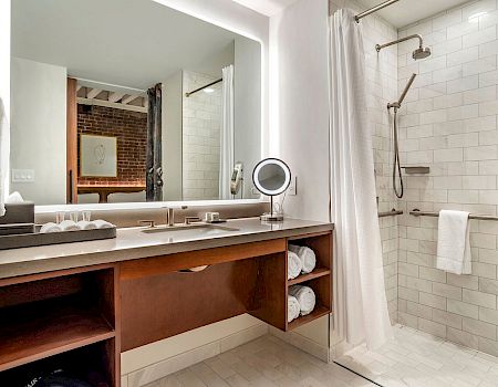 A modern bathroom with a large counter, double sink, open shelves, a lit vanity mirror, and a glass-enclosed shower with white subway tiles, ending this sentence.