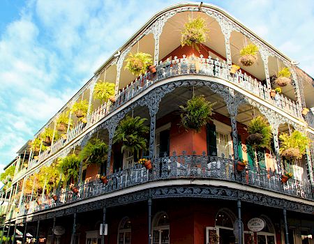 A decorative, multi-story corner building with ornate iron balconies, hanging plants, and bright greenery against a blue sky.