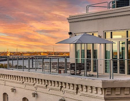 A rooftop terrace at sunset with a glass railing, umbrella, and modern windows overlooking the water, casting warm orange light.