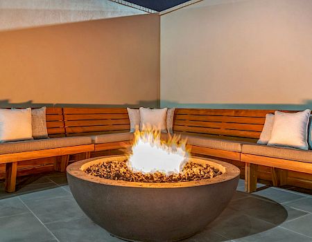 A cozy outdoor seating area with a central fire pit, curved wooden benches, and cushions around warm flickering flames.