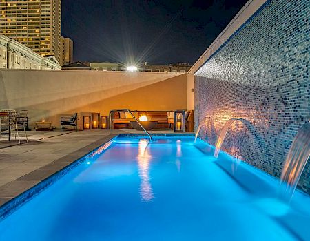 A bright blue rooftop pool with water features and seating under warm lighting at night.