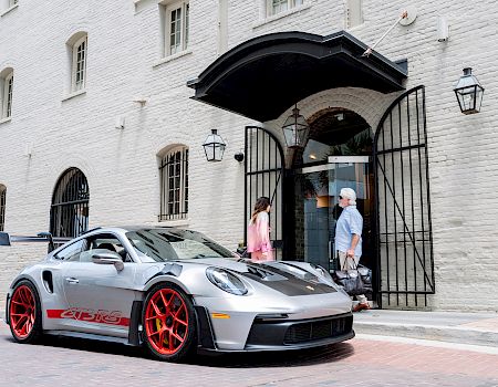 A silver Porsche with red rims parked outside a white brick building; people stand near an open arched doorway.