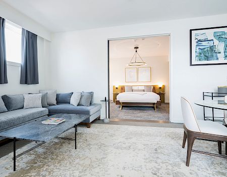 A modern hotel suite with a cozy living area, gray sectional sofa, coffee table, and rug, leading to a bright bedroom with a king bed.