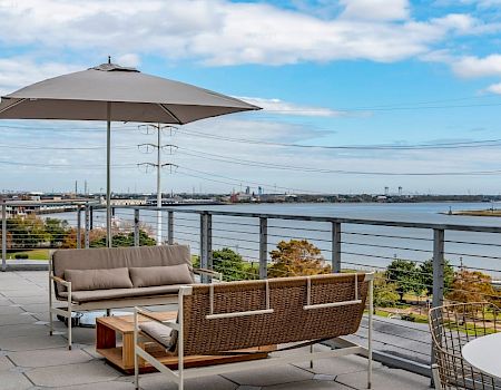 A stylish rooftop terrace with a cushioned seating set, umbrella, and a river view under a bright, partly cloudy sky.