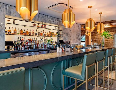 A stylish bar with a long counter, teal seats, gold pendant lights, and shelves of bottles behind the bar.