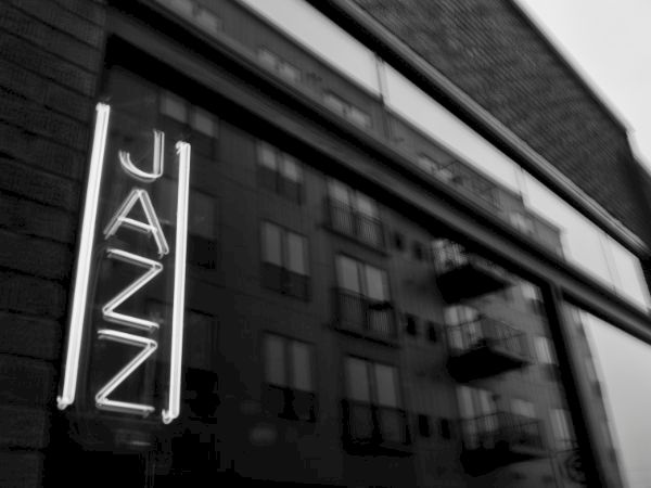 A neon sign reading &ldquo;JAZZ&rdquo; on a building window, reflecting an urban storefront with blurred bricks and windows in the background.