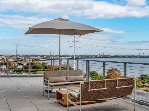 A stylish rooftop outdoor lounge with a cushioned sofa set, umbrella, and a glass railing overlooking a waterway and distant cityscape.