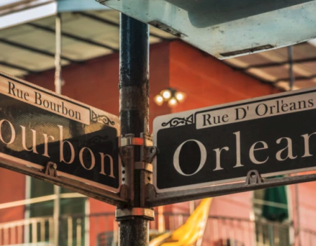 Street signs at a corner show &ldquo;Orleans&rdquo; and &ldquo;Bourbon&rdquo; in a lively downtown scene, with warm building colors and decorative lighting.