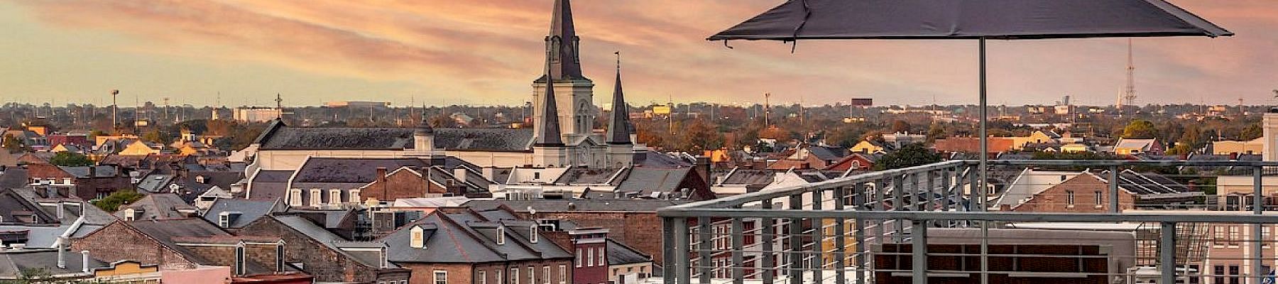 A rooftop deck with a table, umbrella, and chairs overlooks a colorful cityscape at sunset, featuring a church spire and cozy brick buildings.